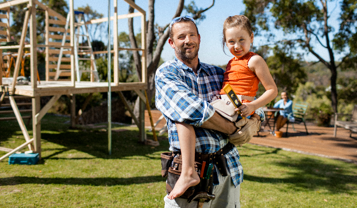 A man in a plaid shirt and tool belt holds a young girl in an orange top, with a construction project in progress behind them in a sunny outdoor setting.