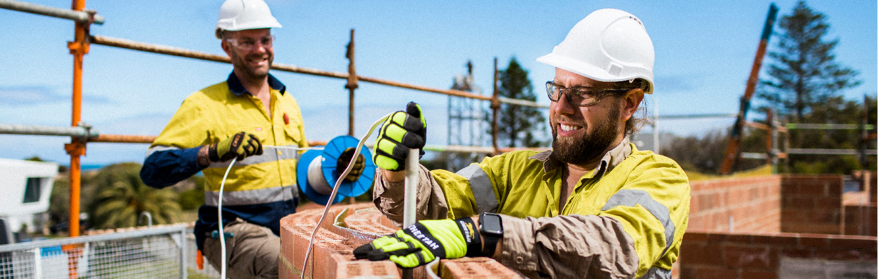 A construction worker in a yellow and navy high-visibility shirt smiles while working on a brick wall, interacting with a colleague on a sunny day at a building site.