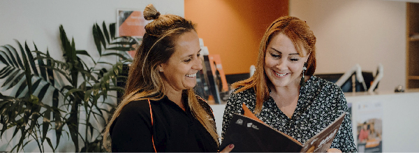 Two young females smiling looking at a brochure together with a palm plant and orange wall behind them