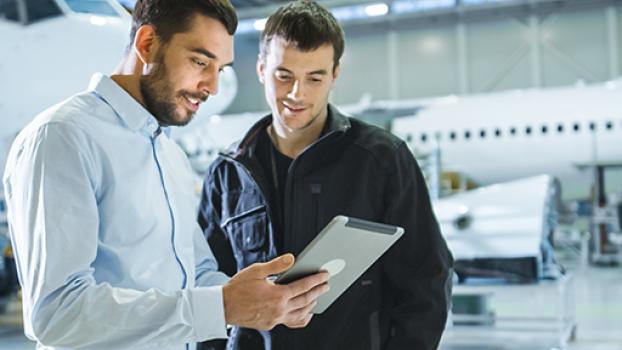 Two male aircraft engineers looking at iPad with planes in a workshop behind them.