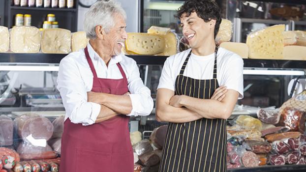 Two smallgoods staff laughing in front of deli counter stocked with meats