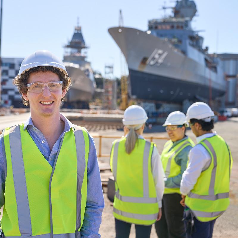 Person in a high-visibility vest and hard hat smiling in a shipyard, with colleagues and large ships in the background.