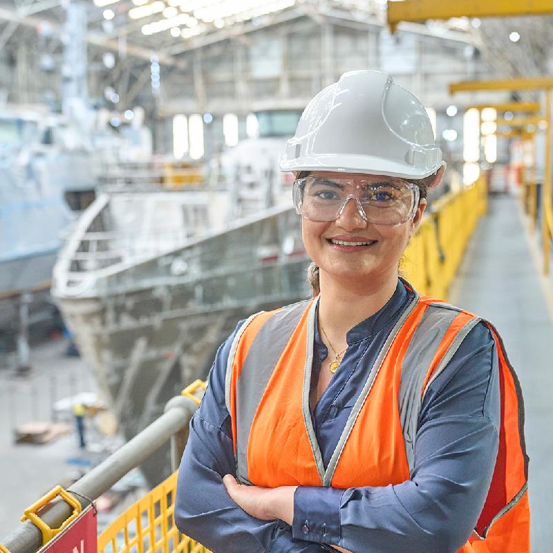 An engineer wearing high visibility protection workwear, stands in front of a marine vessel in a large workshop. 