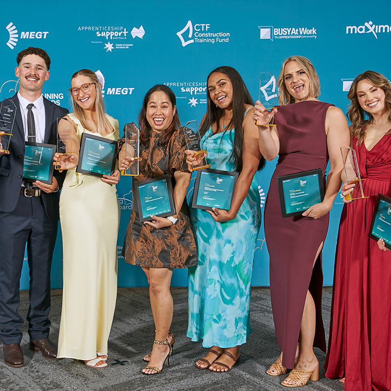 A group of award recipients stand together on a blue event backdrop, each holding a trophy and framed certificate at the WA Training Awards.