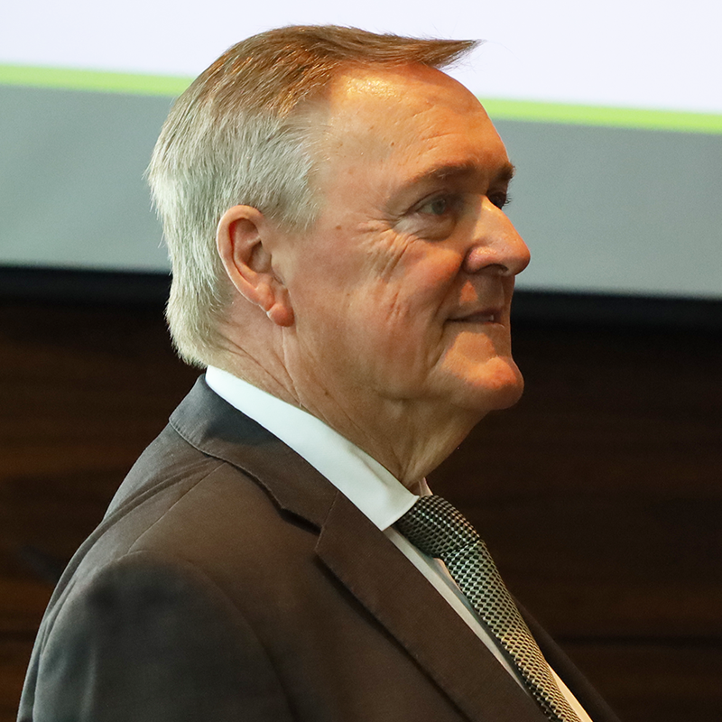 Side profile of a man wearing a dark suit and tie, standing indoors.