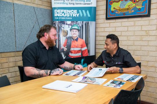 Two people taking at a desk with a defence banner in the background.