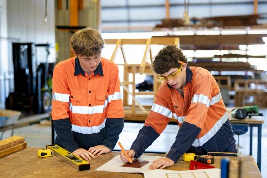 Two male construction students measuring wood on table.