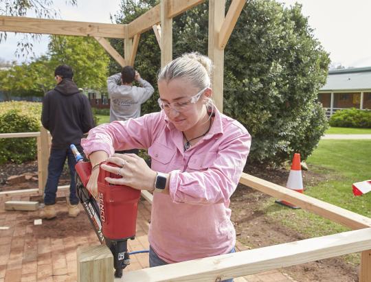 Female construction student using nail gun tool on wood frame.