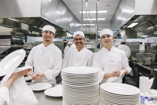 3 chefs smiling at camera in kitchen.